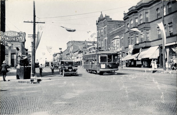 Black and white photo of trolley car, old cars and bicyles driving on a cobblestone street