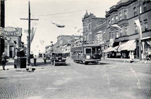 Black and white photo of trolley car, old cars and bicyles driving on a cobblestone street