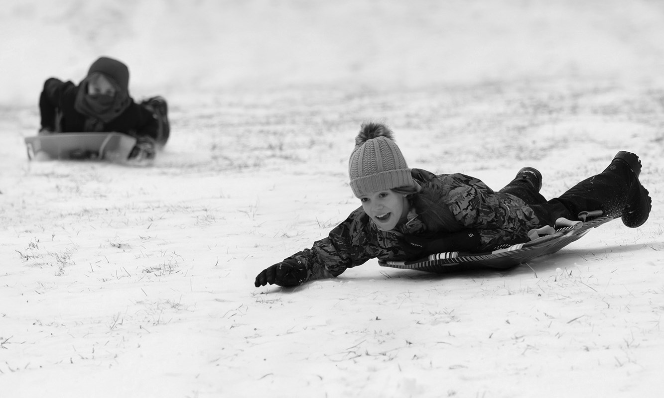 Two kids sledding down a hill on separate sleds, both on their bellies.